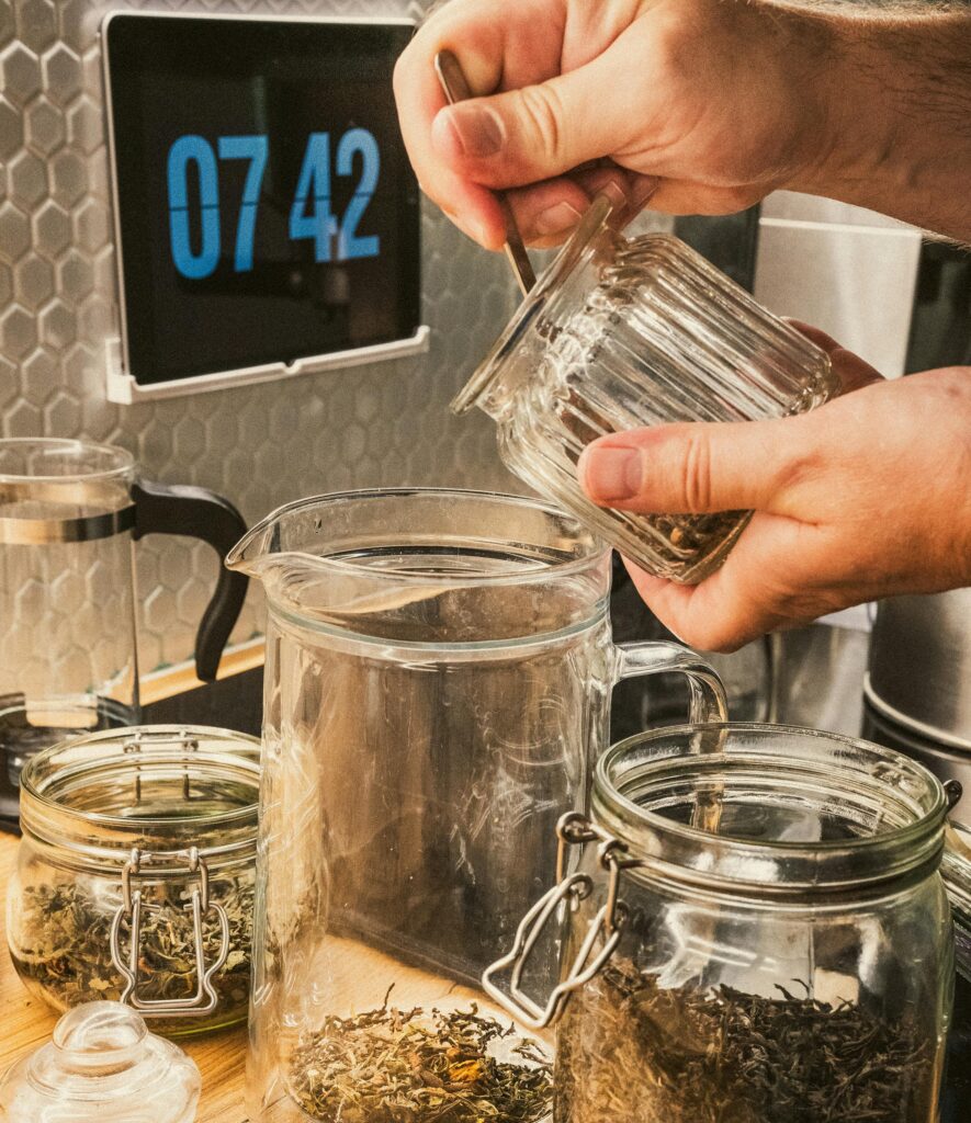 Hands blending loose leaf tea in kitchen setting with glass jars and digital clock display.